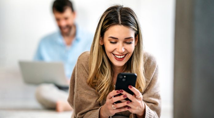 AsianMelodies Review: Check If It’s the Right Communication Platform For You Happy smiling young woman holding smartphone on floor at home using online entertainment mobile app/ AsianMelodies