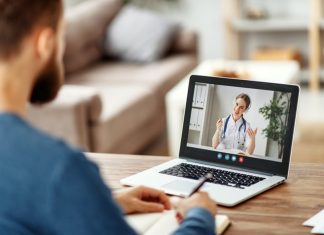 Bearded male patient consulting with female medical practitioner online and writing in notebook while sitting at table at home