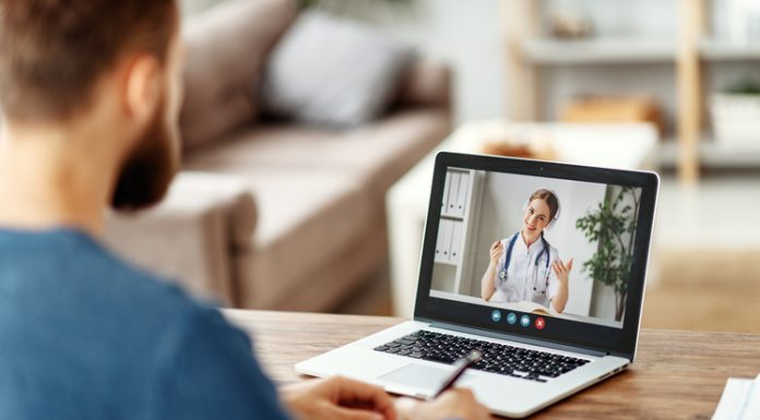 Bearded male patient consulting with female medical practitioner online and writing in notebook while sitting at table at home