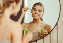 “What if Everything You Knew about Skincare was a Lie?” Says CXC Young woman holding green bottle applying serum to face, looking in mirror, standing in bathroom