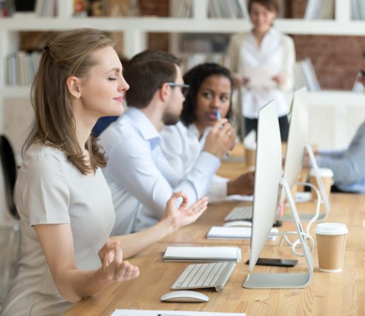 Peaceful millennial woman practice yoga in front of computer at workplace, calm female employee meditating controlling emotions and managing stress, worker sit in lotus position in office