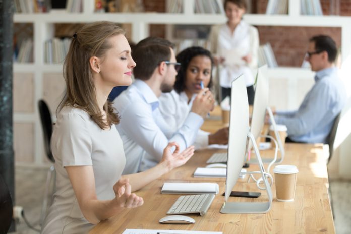 Peaceful millennial woman practice yoga in front of computer at workplace, calm female employee meditating controlling emotions and managing stress, worker sit in lotus position in office