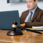 A male lawyer in a brown suit on his computer typing with a gavel front and center on his desk.