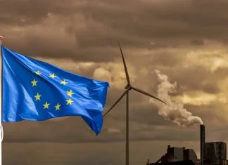 The Role of Policy in a Competitive, More Resilient Europe Waving flag of Europe in front of a polluting factory chimney with smoke and wind turbine