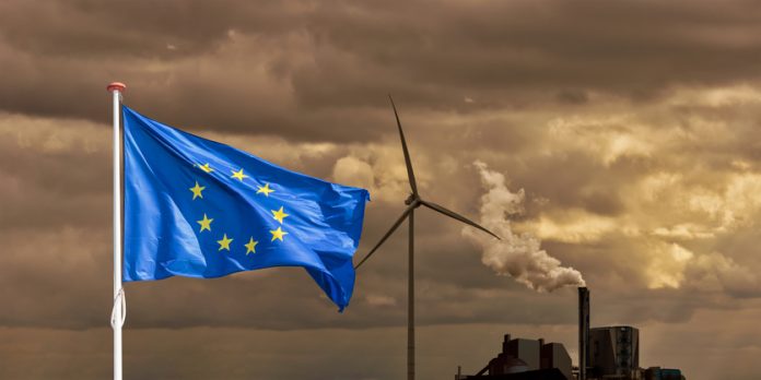 Waving flag of Europe in front of a polluting factory chimney with smoke and wind turbine