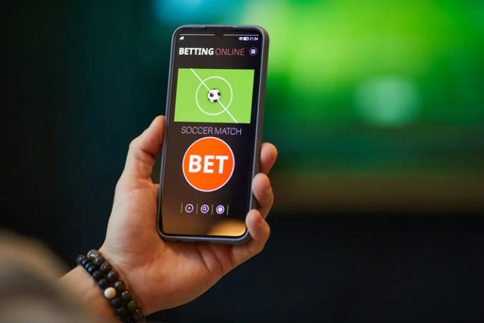 Close-up of hand holding smartphone with soccer betting app interface showing bet options and match information in background
