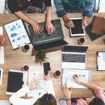 Top view of businessman executive in group meeting with other businessmen and businesswomen in modern office with laptop computer, Playbook, coffee and document on table.