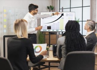 What Influences ERP Software Development Cost: A Complete Guide Team of diverse attentive mixed race business people listening their middle aged male bearded indian chief showing dynamics of key ecological global problems or world market using digital wall screen and ERP software