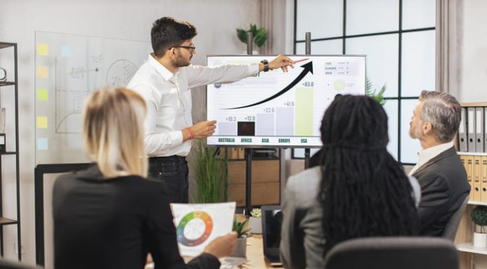 Team of diverse attentive mixed race business people listening their middle aged male bearded indian chief showing dynamics of key ecological global problems or world market using digital wall screen and ERP software