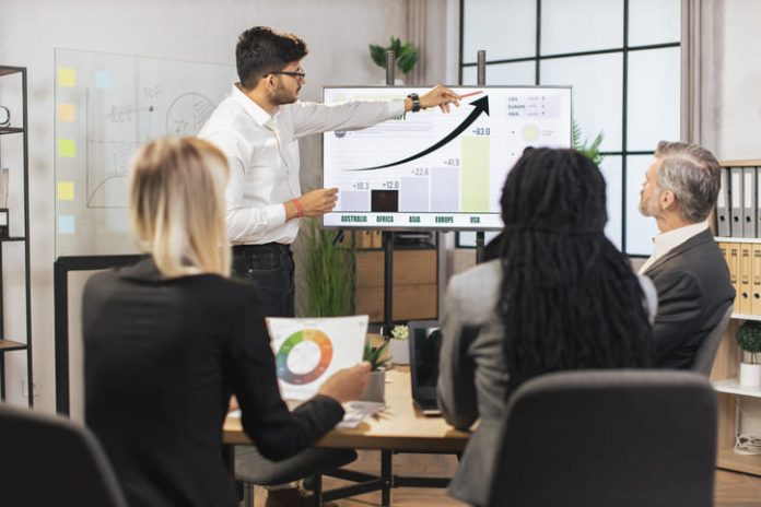 iStock-1392359329 (1) Team of diverse attentive mixed race business people listening their middle aged male bearded indian chief showing dynamics of key ecological global problems or world market using digital wall screen and ERP software