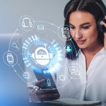 Businesswoman in headphones is holding smartphone with digital interface with various holograms having online conference call. Robot, smart watch, pie diagram. Office workplace with laptop, notebook