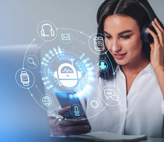 Businesswoman in headphones is holding smartphone with digital interface with various holograms having online conference call. Robot, smart watch, pie diagram. Office workplace with laptop, notebook