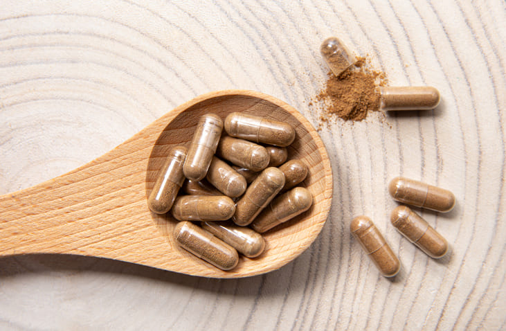 A wooden plate holds a single opened capsule spilling fine brown powder, surrounded by several intact capsules scattered on a marble surface. This image visually represents the premium supplement formulations discussed in the article, symbolizing the scientific and commercial focus on longevity products, such as quercetin-based compounds, within the rapidly growing healthy aging industry.