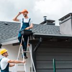 The Value of Certified and Insured Roofers happy certified repairman sitting on roof and holding toolbox near coworker in helmet