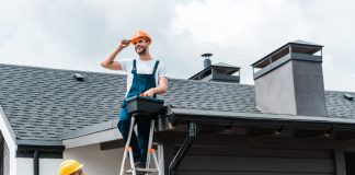 The Value of Certified and Insured Roofers happy certified repairman sitting on roof and holding toolbox near coworker in helmet