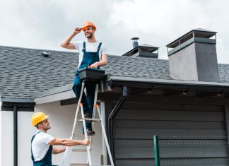 happy certified repairman sitting on roof and holding toolbox near coworker in helmet