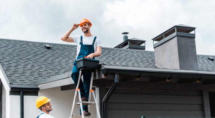 The Value of Certified and Insured Roofers happy certified repairman sitting on roof and holding toolbox near coworker in helmet
