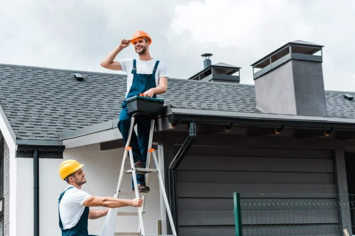 happy certified repairman sitting on roof and holding toolbox near coworker in helmet