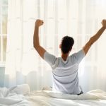 The Power of Self-Care in Boosting Your Productivity Guy sitting on bed facing the window, awakening after good sleep and rest prioritizing self-care