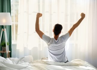 Guy sitting on bed facing the window, awakening after good sleep and rest prioritizing self-care