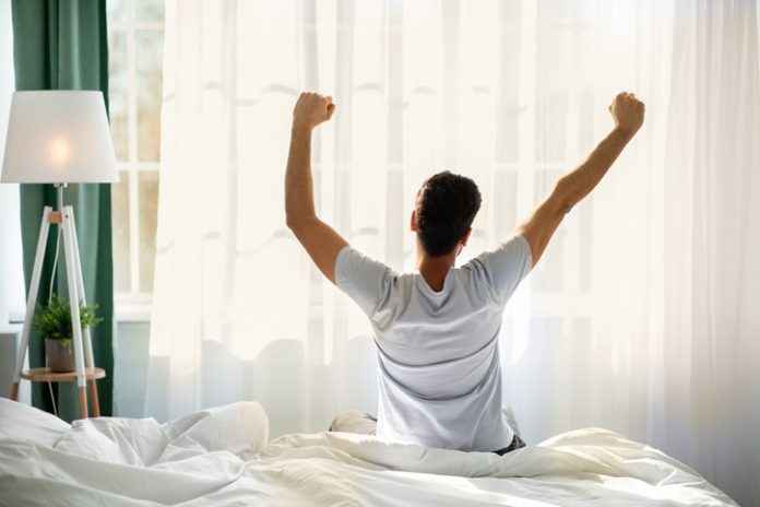 Guy sitting on bed facing the window, awakening after good sleep and rest prioritizing self-care