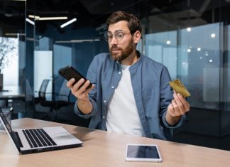 Frustrated and deceived businessman in office trying to make purchase in online store and online money transfer from bank credit card, annoyed man working inside office using laptop and phone. Sugar baby scam online