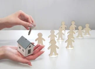 Hand holding a model of a house and a key, offering it to figures of people. Supportive housing concept