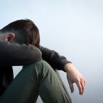 Why Mental Health Is Suffering in the UK Person sitting in low light holding their legs, symbolizing stress and anxiety caused by the cost of living and the growing mental health crisis in the UK.