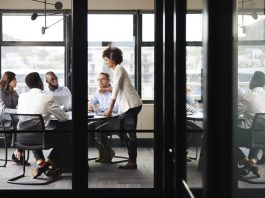 Millennial businesswoman stands addressing colleagues and thinking new strategy at a meeting, seen through glass wall
