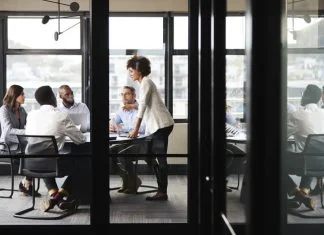 Millennial businesswoman stands addressing colleagues and thinking new strategy at a meeting, seen through glass wall