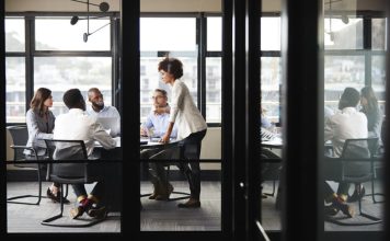 Millennial businesswoman stands addressing colleagues and thinking new strategy at a meeting, seen through glass wall