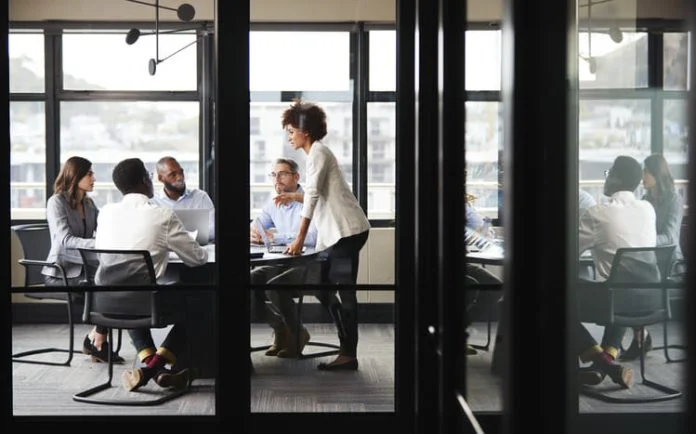 iStock-1146473085 (1) Millennial businesswoman stands addressing colleagues and thinking new strategy at a meeting, seen through glass wall