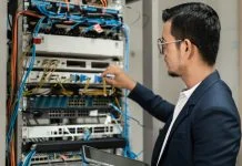 Young network technician holding tablet working to connecting network cables in server cabinet in network server room.