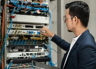 Young network technician holding tablet working to connecting network cables in server cabinet in network server room.