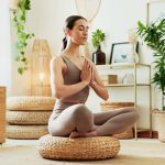 A woman meditating in a bright living room surrounded by indoor plants, practicing mindfulness, wellness and relaxation.
