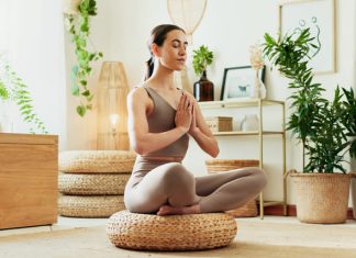 A woman meditating in a bright living room surrounded by indoor plants, practicing mindfulness, wellness and relaxation.
