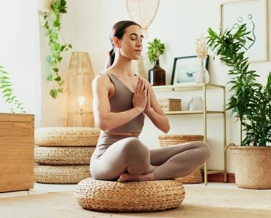A woman meditating in a bright living room surrounded by indoor plants, practicing mindfulness, wellness and relaxation.