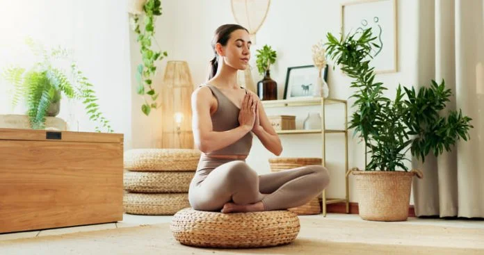 iStock-2177247670 (1) A woman meditating in a bright living room surrounded by indoor plants, practicing mindfulness, wellness and relaxation.