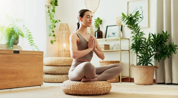 From Burnout to Balance: How Recovery Principles Are Shaping the Future of Workplace Wellness A woman meditating in a bright living room surrounded by indoor plants, practicing mindfulness, wellness and relaxation.