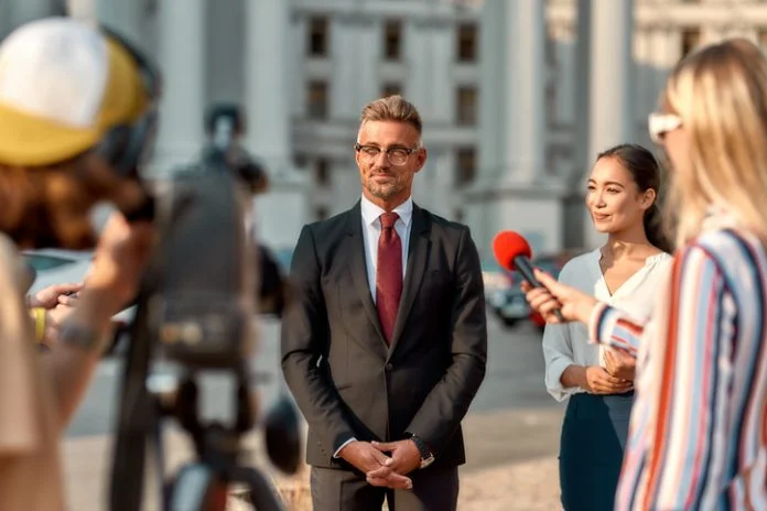 Horizontal shot of politician giving interview during campaign to journalists. People making interview using digital camera and equipment set at outdoor location.