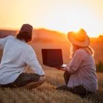 Farmers using laptop for smart farming analysis while observing rural field during sunset. Concept of modern agriculture and farm innovation