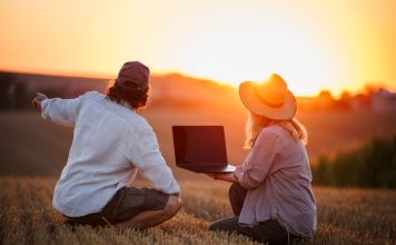 Farmers using laptop for smart farming analysis while observing rural field during sunset. Concept of modern agriculture and farm innovation