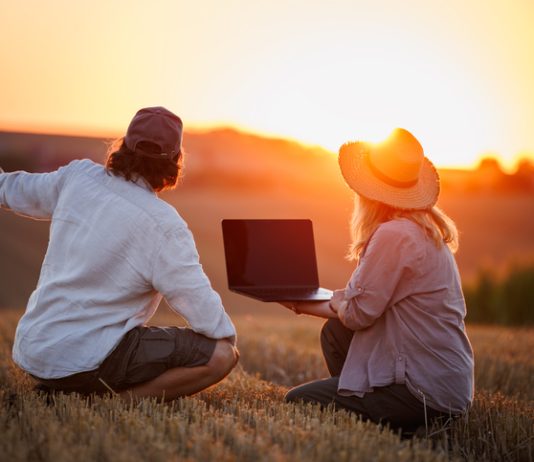 Farmers using laptop for smart farming analysis while observing rural field during sunset. Concept of modern agriculture and farm innovation