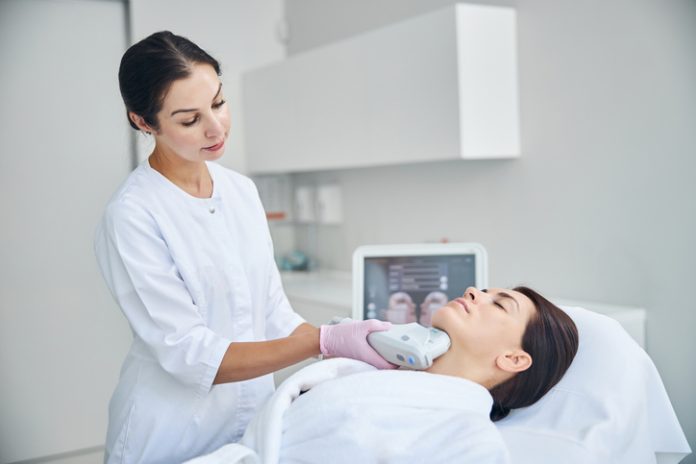 Attractive lady lying still during the ultrasound therapy Experienced female dermatologist using a Ultherapy Prime beauty device for tightening the skin on her patient neck