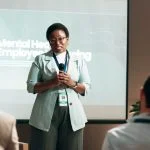 Seminar, speaker and woman with mic, talking and public speaking for mental health in workplace or stage. Business, presentation and employee with speech for audience, people and podium for presenter