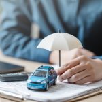 The Real Cost of a Car: Why Insurance Should Influence Your Choice Car insurance protect concept. Insurance agent. Insurance protection concept. A person is holding a blue car under an umbrella. The umbrella is white and is placed on top of a piece of paper.