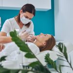 Brunette beautician in uniform holding syringe with an injection needle for Juvederm Voluma in Clinical Practice while patient lying on medicine chair in beauty salon