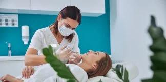 Brunette beautician in uniform holding syringe with an injection needle for Juvederm Voluma in Clinical Practice while patient lying on medicine chair in beauty salon