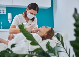 Brunette beautician in uniform holding syringe with an injection needle for Juvederm Voluma in Clinical Practice while patient lying on medicine chair in beauty salon
