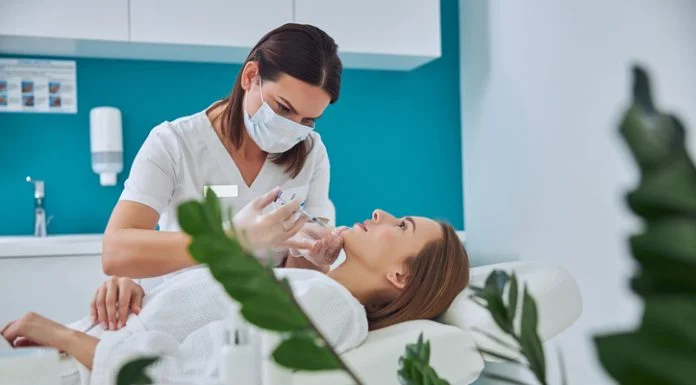 Brunette beautician in uniform holding syringe with an injection needle for Juvederm Voluma in Clinical Practice while patient lying on medicine chair in beauty salon